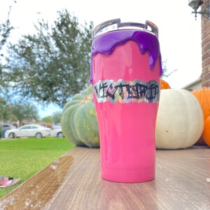 Puede incluir: Un vaso rosa con un dise&ntilde;o de goteo morado y el nombre "VICTORIA" en letras plateadas. El vaso est&aacute; sobre una superficie de madera con calabazas en el fondo.