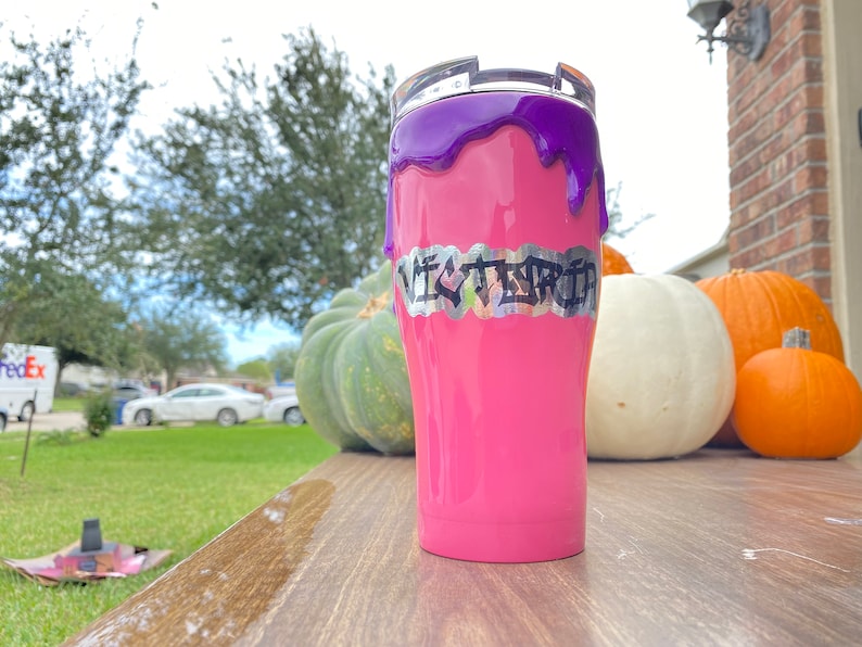 Puede incluir: Un vaso rosa con un dise&ntilde;o de goteo morado y el nombre "VICTORIA" en letras plateadas. El vaso est&aacute; sobre una superficie de madera con calabazas en el fondo.