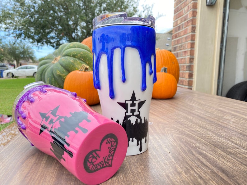 Puede incluir: Dos vasos aislados con base blanca y dise&ntilde;o de pintura goteada azul o rosa. Un vaso tiene un dise&ntilde;o de pintura goteada azul, y el otro tiene un dise&ntilde;o de pintura goteada rosa. Ambos vasos tienen un gr&aacute;fico negro de una estrella y un horizonte.