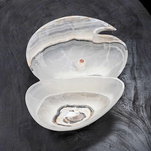 May include: A carved onyx bowl with a unique, organic shape. The bowl features a light, translucent color with gray and brown veining. The design includes a top section with a curved opening and a lower, rounded bowl. The overall dimensions are approximately 8 inches wide.