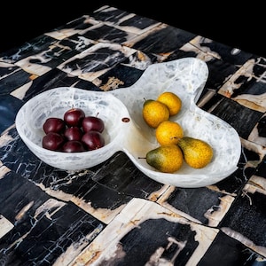 May include: A white, three-section serving dish filled with fresh fruit. The left section holds dark red plums, while the center and right sections contain yellow pears. The dish sits on a black and white patterned surface.