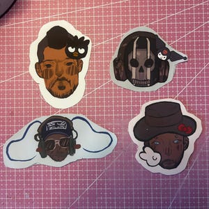 May include: Four die-cut stickers featuring illustrated faces. One has a black bird, another a skull with headphones, a third with sunglasses and a hat, and the last with a hat and a cloud-shaped design. The stickers are on a pink grid surface.