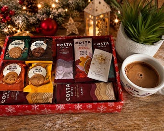 Może przedstawiać: Czerwone pudełko prezentowe zawierające różne saszetki Costa Coffee Barista Creations oraz paczki ciastek, w tym smaki Golden Oat, Belgian Chocolate, Salted Caramel i Stem Ginger. Obok pudełka stoi biały ceramiczny kubek wypełniony kawą.