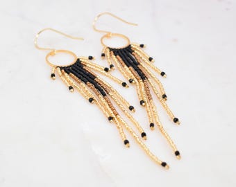 May include: A pair of gold-tone beaded earrings with a fringe design. The earrings feature a gold hoop and are adorned with black, gold, and bronze-colored beads. The earrings are displayed on a white surface.
