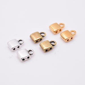 Puede incluir: Una colección de hallazgos de joyería de metal en tonos plateados, dorados y oro antiguo. Los hallazgos son pequeños, rectangulares con un lazo y dos agujeros, adecuados para la fabricación de joyas. Los hallazgos están dispersos sobre una superficie blanca.