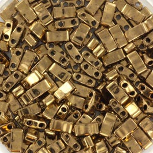 Könnte beinhalten: Nahaufnahme eines Haufens kleiner, rechteckiger, goldfarbener Perlen. Jede Perle hat zwei Löcher zum Auffädeln. Die Perlen sind dicht gepackt, reflektieren das Licht und erzeugen einen schimmernden Effekt. Ideal für die Schmuckherstellung.