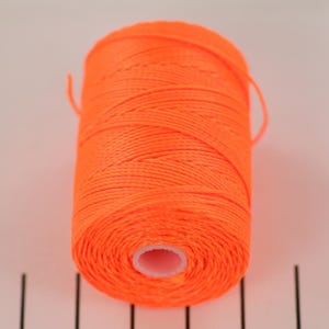 Könnte beinhalten: Eine Spule leuchtend orangefarbenen Fadens. Der Faden ist eng um einen weißen Kern gewickelt und bildet eine zylindrische Form. Der Faden scheint aus einem synthetischen Material zu bestehen.