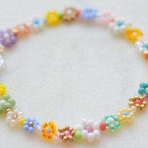 A zoom up of a bracelet made with seed beads of all colors and sizes. The beads are arranged in little flowers. Each flower is different and there is also a different bead between each flower. The clasp is gold.