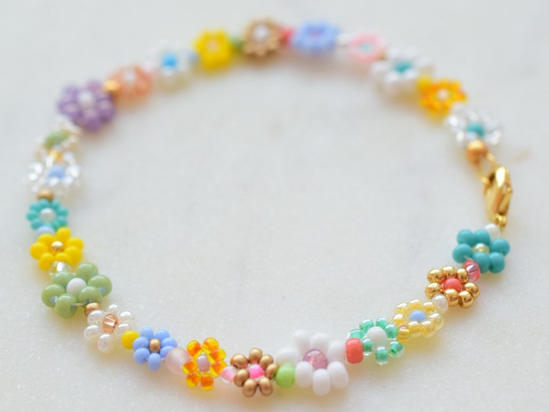 A zoom up of a bracelet made with seed beads of all colors and sizes. The beads are arranged in little flowers. Each flower is different and there is also a different bead between each flower. The clasp is gold.