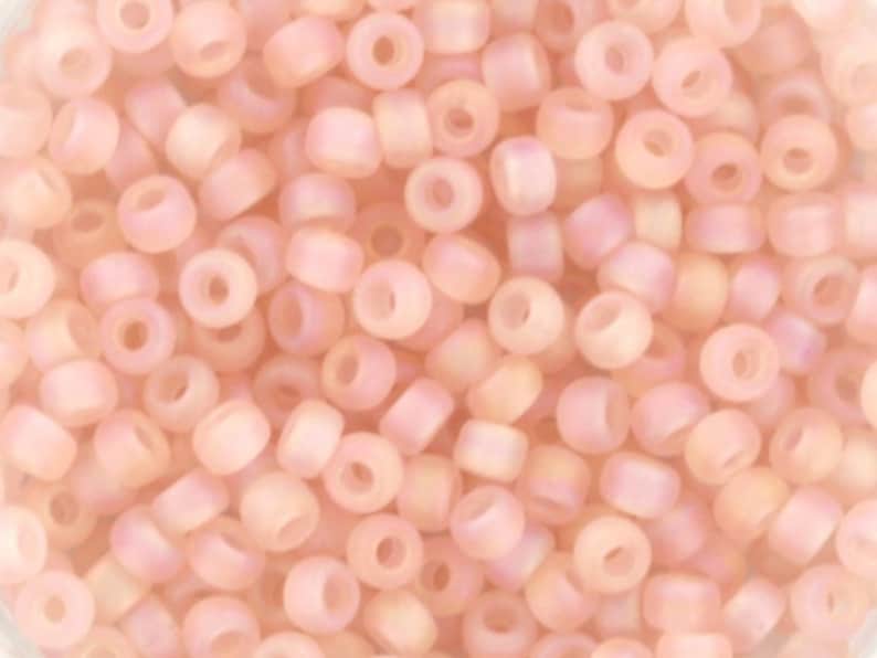 Pu&ograve; includere: Una vista ravvicinata di un mucchio di piccole perline rotonde di colore rosa chiaro con un sottile bagliore iridescente. Le perline sono opache e hanno una finitura liscia e lucida.
