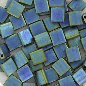 Könnte beinhalten: Eine Ansammlung quadratischer, irisierender blauer Perlen mit metallischem Glanz. Die Perlen sind verstreut und zeigen Farbvariationen von Türkis bis Tiefblau. Jede Perle hat ein kleines Loch zum Auffädeln.