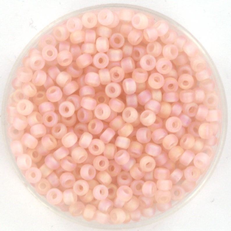 Pu&ograve; includere: Una vista ravvicinata di una ciotola piena di piccole perline rotonde, opache e rosa. Le perline hanno una finitura leggermente iridescente, che conferisce loro un leggero bagliore.