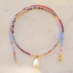 Op de afbeelding: Een delicate ketting met een parel hanger. De ketting is gemaakt van kleine kralen in tinten roze, paars, blauw en goud.
