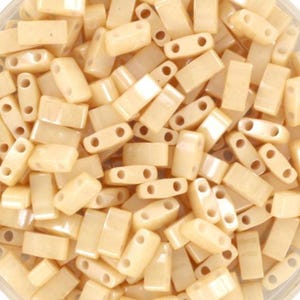 Puede incluir: Primer plano de un montón de cuentas rectangulares de color beige claro. Cada cuenta tiene dos agujeros para ensartar. Las cuentas son lisas y tienen un aspecto ligeramente translúcido, lo que sugiere que están hechas de vidrio o un material similar. Están dispersas al azar.