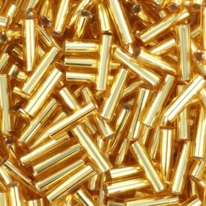 Puede incluir: Una imagen de primer plano de un montón de cuentas de vidrio doradas. Las cuentas son largas y cilíndricas, con un acabado brillante y metálico.