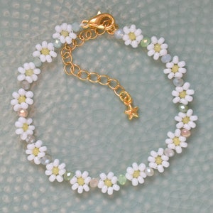 Könnte beinhalten: Ein zartes Armband aus weißen Perlen in Form von Gänseblümchen mit einer goldenen Kette und einem Sternanhänger. Das Armband besteht aus kleinen weißen Perlen, die die Form von Gänseblümchen haben.