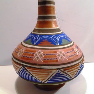 Puede incluir: Un jarrón de cerámica pintado a mano con un cuerpo bulboso y un cuello estrecho. El jarrón presenta una paleta de colores marrón, naranja, rosa, azul y blanco. Patrones geométricos y bandas decorativas adornan la superficie. El jarrón mide unos 25 cm de altura.