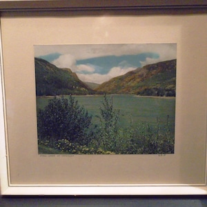 May include: Framed landscape print featuring a serene lake scene surrounded by mountains and lush greenery. The artwork is set within a white frame, enhancing the natural colors of the landscape. The print includes text at the bottom.