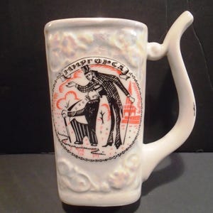 May include: White ceramic mug with a unique handle design. The mug features a black and red illustration of two figures in formal attire within a circular frame. The mug has a textured surface and the text "МИРГОРСА" is visible.