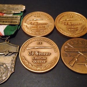 May include: A collection of vintage shooting medals and awards. The medals are bronze-colored and feature inscriptions such as "To Honour" and "B.C. Rifle Association." One medal has a ribbon with green, white, and orange stripes. The awards are from 1901 to 1961.