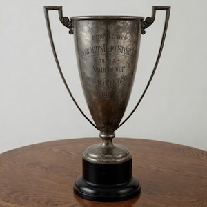 May include: A silver trophy with a black base, featuring handles and engraved text. The text reads "DONATED BY HOWARD'S DEPT STORE TO THE Vancouver Homing Pigeon Society 1927". The trophy is displayed on a wooden table.