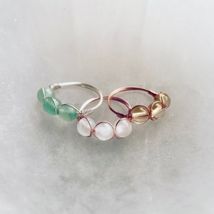 May include: Three wire-wrapped rings with different colored stones. The first ring has three green stones, the second ring has three white stones, and the third ring has three yellow stones.