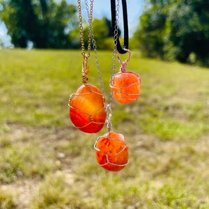 May include: Three carnelian stone necklaces with wire wrapped pendants. The stones are orange and red with a translucent quality. The necklaces are on a white background.