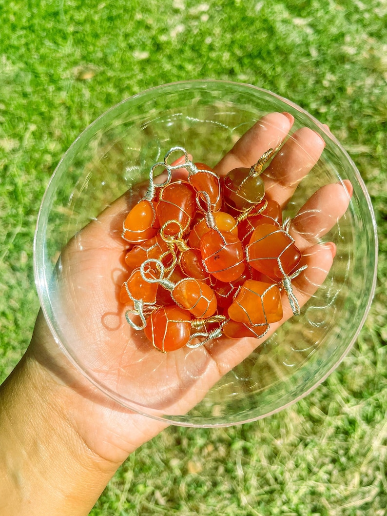 May include: A collection of carnelian stones wrapped in gold wire. The stones are a vibrant orange color and are displayed in a clear glass bowl.