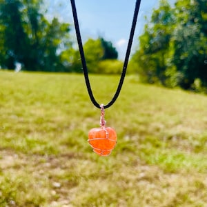 May include: A delicate necklace with a peach-colored stone wrapped in copper wire. The necklace is hanging against a blurred background of green grass and trees.