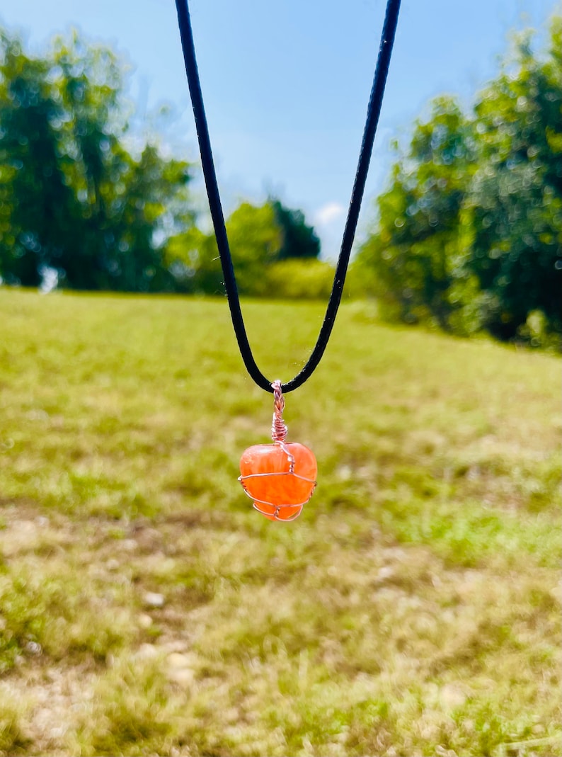 May include: A delicate necklace with a peach-colored stone wrapped in copper wire. The necklace is hanging against a blurred background of green grass and trees.