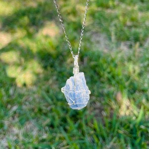 K&ouml;nnte beinhalten: Eine silberne Kettenkette mit einem blauen Kristallanh&auml;nger, der in Silberdraht gewickelt ist. Der Anh&auml;nger ist ein roher, nat&uuml;rlicher Kristall in hellblauer Farbe.