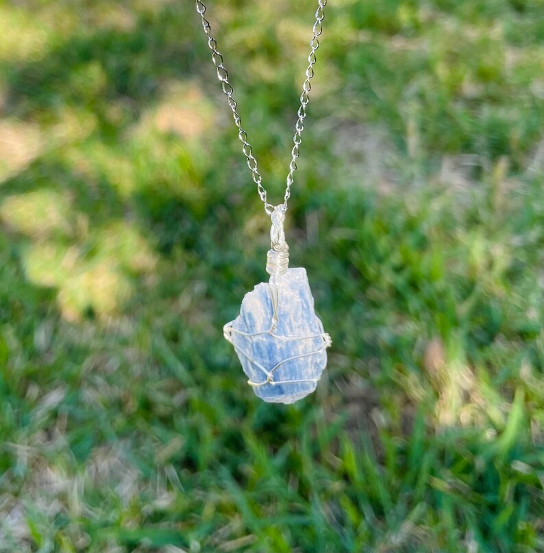 K&ouml;nnte beinhalten: Eine silberne Kettenkette mit einem blauen Kristallanh&auml;nger, der in Silberdraht gewickelt ist. Der Anh&auml;nger ist ein roher, nat&uuml;rlicher Kristall in hellblauer Farbe.