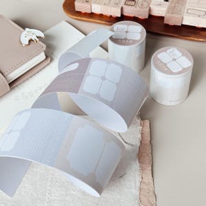 以下が含まれることがあります： チケットデザインの装飾テープ、木製スタンプ、小さなノートなど、文房具のコレクション。テープは薄いグレーで、繰り返し模様があり、スタンプは木製のトレイに入っています。