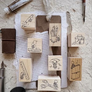 May include: A collection of wooden stamps with black ink illustrations. The stamps feature various designs, including a pen, pencils, a ruler, scissors, and a book, each with a whimsical character. A brown leather journal and a tube of paint are also visible.