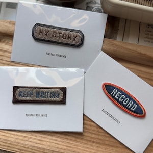 Puede incluir: Tres parches bordados sobre tarjetas blancas. Uno dice "MY STORY" en hilo marrón, otro "KEEP WRITING" y el tercero "RECORD". Los parches están sobre una bandeja de madera con otros artículos de papelería.
