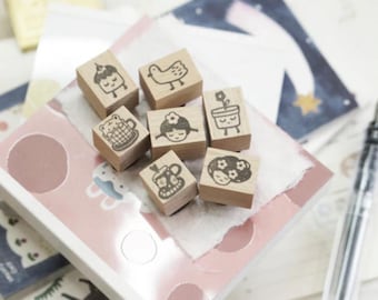 May include: A collection of seven small, wooden stamps with various cute illustrations, including a cupcake, bird, bear in a teacup, girl, flower pot, skull with a mug, and a girl with flowers. The stamps are arranged on a pink and white patterned surface.