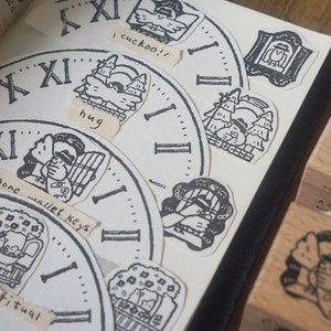 May include: A collection of black and white rubber stamps featuring various illustrations, including a clock face, a bird, a cat, and a person holding a phone. The stamps are on a white background with handwritten text that reads "I cuckoo!!", "hug", "phone wallet keys", and "A sense of ritual (open)".