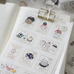 Può includere: Fogli di adesivi decorativi con illustrazioni stravaganti in un design di francobollo. Gli adesivi presentano simpatici personaggi, cibi e oggetti, con dettagli dorati e testo scritto a mano. Gli adesivi sono tenuti in posizione con una clip di metallo.