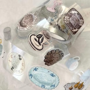 Op de afbeelding: Een rol transparante washi tape met aquarel illustraties van kopjes, borden en serveerschalen. De ontwerpen omvatten bloemmotieven en een zacht kleurenpalet van blauw, bruin en roze. Ideaal voor scrapbooking en knutselen.