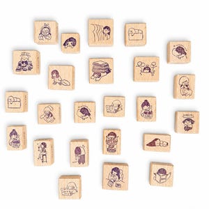 Op de afbeelding: Een verzameling van tweeëntwintig vierkante en rechthoekige houten stempels, elk voorzien van een unieke donkerpaarse inktillustratie van een stripfiguur tijdens diverse dagelijkse activiteiten, zoals lezen, koffiedrinken en uitrusten.