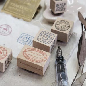 May include: A collection of wooden stamps with various designs and text, including a stamp with a cake and the words "This Moment, Cherished in Memory". A fountain pen and stamped paper are also visible.