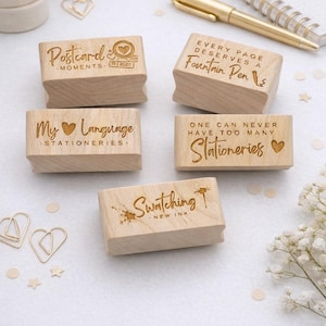 Peut inclure: Tampons en bois avec des inscriptions gravées, dont "Postcard Moments 2020", "Every Page Deserves a Fountain Pen", et "My Language Stationeries". Les tampons sont rectangulaires et en bois clair.