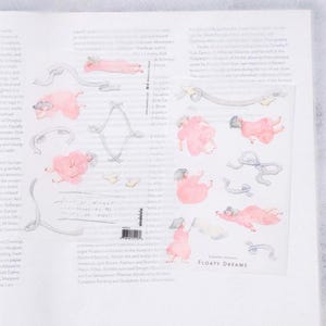 May include: A sheet of transfer stickers featuring watercolor illustrations of figures in pink dresses and gray hair, floating with gray ribbons. The text "FLOATING DREAMS" is printed in black.