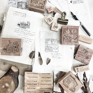 May include: A collection of wooden stamps, stationery, and handwritten notes. The stamps feature illustrations of a typewriter, a bear, and other objects. The text "PAGES OF ME" is visible on a notebook. The items are arranged on a white surface.