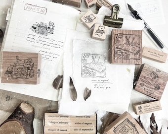 May include: A collection of wooden stamps, stationery, and handwritten notes. The stamps feature illustrations of a typewriter, a bear, and other objects. The text "PAGES OF ME" is visible on a notebook. The items are arranged on a white surface.