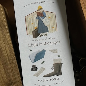 Peut inclure: Une impression décorative sur papier avec le texte "note paper" et "Light in the paper". L'illustration représente une personne lisant un livre, une trousse, un flacon d'encre et une botte. Le texte "YAMADORO" est également présent.
