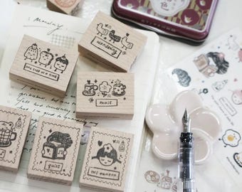 May include: A collection of wooden stamps with whimsical illustrations and text, including images of birds, food, and cute characters. The stamps are arranged on a white surface with a pen and stickers, perfect for journaling and scrapbooking.