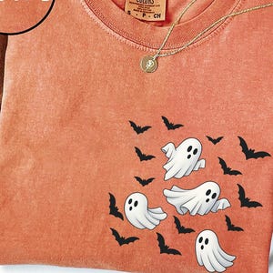 May include: Orange Comfort Colors t-shirt featuring a design of cartoon ghosts and bats. The shirt has a round neck and short sleeves. A gold necklace with a pendant is draped across the shirt. A brown hat is in the background.