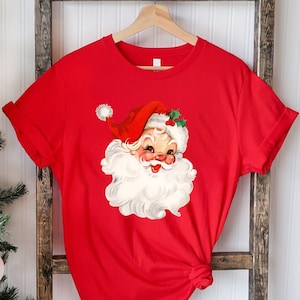 May include: A red t-shirt featuring a vintage illustration of Santa Claus with a white beard, red hat, and rosy cheeks. The Santa Claus graphic is centered on the shirt. The shirt is hanging on a wooden hanger against a rustic wooden ladder.