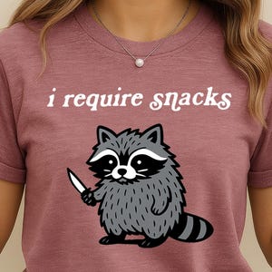 May include: Mauve-colored t-shirt with the text "i require snacks" above a cartoon raccoon holding a knife. The raccoon is gray and white with a black and white striped tail. The shirt is a crew neck.